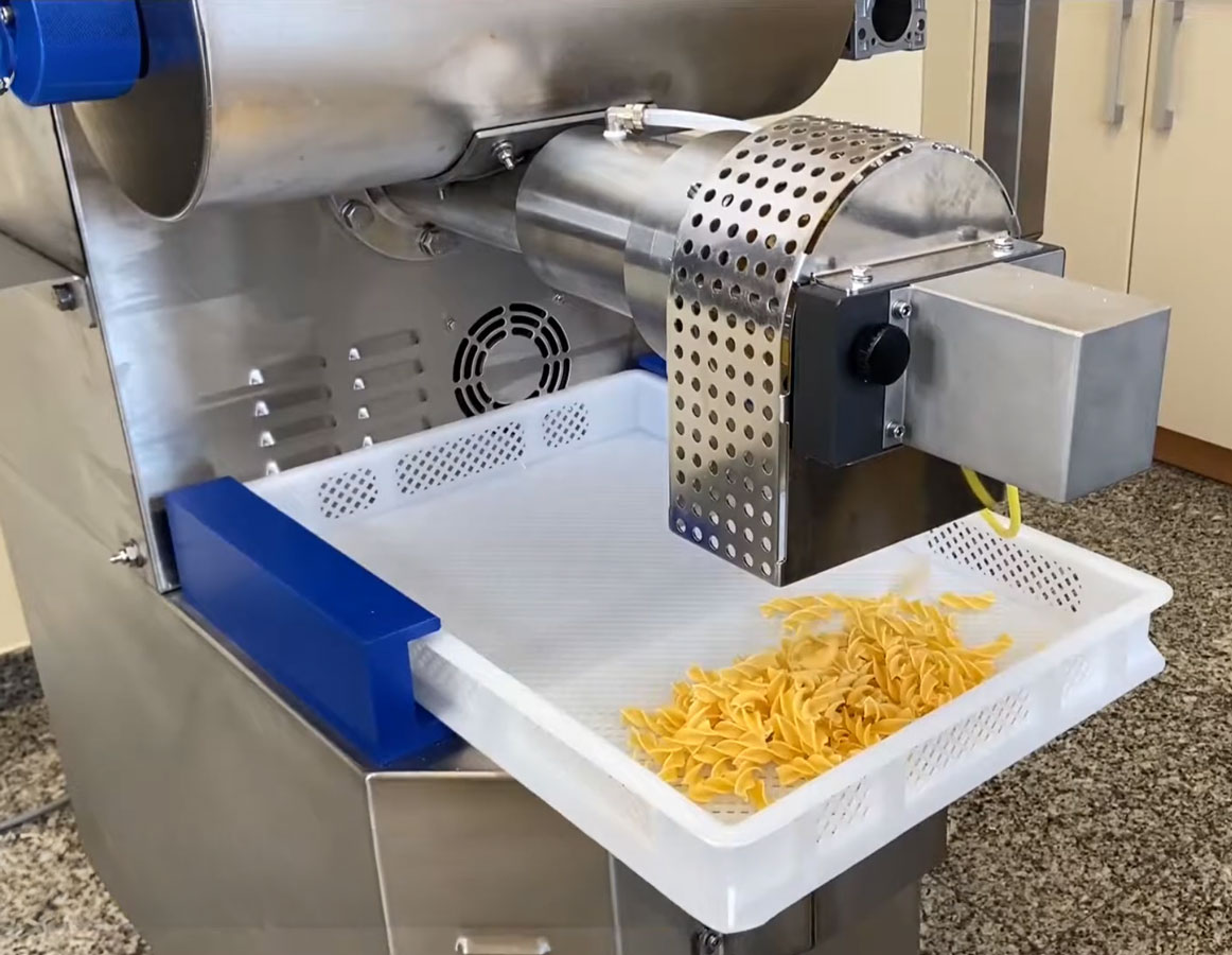 A pasta machine extruding spiral-shaped noodles into a white pasta basket.